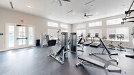 Located at 2213 Highland Forest Dr in Richmond, VA, this dedicated fitness center offers a generously sized, single-floor workout space designed to accommodate a full range of exercise needs. The facility is bathed in natural light streaming through a series of large, elevated windows fitted with blinds, creating a bright and energizing atmosphere throughout.

The space is thoughtfully organized into distinct workout zones. A strength training area features a multi-function cable machine, a power rack with weight storage, a full dumbbell rack, and adjustable benches — providing everything needed for a comprehensive resistance training session. Large wall-mounted mirrors enhance the sense of space and allow users to monitor their form.

The cardio section is equally well-appointed, housing multiple treadmills, an elliptical, and a stair climber, all from the Matrix brand. Ceiling fans positioned overhead ensure consistent airflow and comfort during intense workouts.

The flooring throughout is durable dark rubber, ideal for both heavy equipment and high-impact activities. Recessed lighting supplements the abundant natural light, while a direct exterior door provides convenient access. The clean, neutral color palette and modern finishes give the space a professional, welcoming ambiance suited for everyday fitness use.