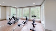 The Spin Room is a dedicated fitness space designed to support focused cardio workouts in a bright, comfortable environment. The room is finished with smooth hardwood-style flooring that runs throughout, providing a clean and durable surface well-suited for exercise equipment and open floor activities. Neutral-toned walls with gray crown molding contribute to a calm, distraction-free atmosphere. Large multi-pane windows line multiple walls, flooding the space with abundant natural light while offering expansive views of lush greenery and surrounding landscaping — creating an energizing, nature-connected backdrop for workouts. The room is equipped with spin bikes and rowing machines, making it versatile for a range of cardio-focused exercise. Recessed lighting and a ceiling fan provide consistent overhead illumination and airflow, ensuring comfort during intense sessions. A decorative column with vertical slat detailing adds an architectural accent near the entry, lending the space a refined, intentional design. The open floor plan allows for flexible arrangement of equipment or additional fitness activities, while the overall layout promotes a focused yet inviting workout environment. This well-lit, thoughtfully designed spin room balances function and aesthetics for an elevated fitness experience.