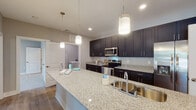 Located at 4525 Croquet Dr in Wilmington, NC, this single-story home offers a thoughtfully designed open-concept layout with contemporary finishes throughout. The kitchen is a standout feature, showcasing dark espresso shaker-style cabinetry, granite countertops, a large center island with an undermount sink, pendant lighting, and a full suite of stainless steel appliances including a range, microwave, and French door refrigerator. The open living area flows seamlessly from the kitchen, featuring durable wood-look flooring, neutral-toned walls, recessed lighting, and direct access to an outdoor area through glass doors that invite natural light. The bedrooms are carpeted for comfort, with large windows framing views of surrounding greenery. The primary bedroom includes access to a walk-in closet and an en-suite bath featuring granite countertops and a glass-enclosed shower. A dedicated laundry room houses a full-size washer and dryer. The bathrooms continue the home's cohesive design with matching dark cabinetry and granite surfaces. Clean lines, neutral tones, and quality materials create a modern yet welcoming atmosphere throughout this well-appointed Wilmington home.
