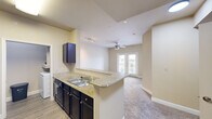Located at 1300 Placid Cir in Waco, TX, this one-bedroom, two-bathroom unit offers a well-planned layout across a single floor. The galley-style kitchen is a functional centerpiece, featuring dark cabinetry with brushed hardware, granite-style countertops, a double-basin sink, and a full suite of black appliances including a refrigerator, electric range, microwave, and dishwasher. Wood-look flooring runs throughout the kitchen, which opens toward the living area and connects directly to an in-unit laundry room. The living area is finished with plush carpet and is brightened by natural light streaming through French doors that lead to a private balcony. A ceiling fan with light fixture adds comfort to the space. The bedroom offers generous proportions with carpet flooring, a large window with blinds for natural light, a ceiling fan, and direct access to a dedicated walk-in closet equipped with built-in shelving. The two bathrooms are well-appointed—one features a sliding glass-door shower with tile surround and chrome fixtures, while the other includes a wide vanity with dark cabinetry, granite-style counters, and wood-look flooring. Neutral warm-toned walls and white trim throughout give the space a clean, cohesive aesthetic.