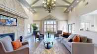 Located at 5225 Zachary Grove in Colorado Springs, CO, this single-story community clubhouse delivers an impressive blend of architectural character and practical amenities. The grand entrance sets the tone immediately, framed by stacked stone columns, arched alcoves with potted plants, and exposed brick accents that open into a soaring great room. Vaulted ceilings with decorative beams, an elegant chandelier, and a wall of large windows flood the lounge with natural light, while plush seating arrangements around a glass coffee table create a welcoming social hub. A stone fireplace surround anchors one wall, adding warmth and texture to the space. Adjacent to the lounge, a well-equipped kitchen bar features granite countertops, stainless steel appliances, and bar stool seating—ideal for casual gatherings. A separate dining area provides communal bench seating at a solid wood table beneath track lighting, with direct outdoor access. The spacious fitness center is outfitted with a comprehensive selection of cardio machines, strength equipment, ceiling fans, wall mirrors, and mounted televisions, all set on durable wood-look flooring. A dedicated business office rounds out the facility, offering a quiet workspace with a round table and artwork. Throughout, the design balances rustic stone detailing with clean, modern finishes.