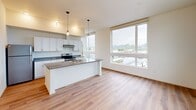Located on the fifth floor at 310 Capitol Way N in downtown Olympia, Washington, this contemporary residence offers a bright and thoughtfully designed single-level layout. The open-concept living and kitchen area serves as the heart of the unit, featuring warm hardwood-style flooring that flows seamlessly throughout the space. The kitchen is outfitted with crisp white cabinetry, dark countertops, stainless steel appliances including a refrigerator and electric range, and a central island with pendant lighting overhead—ideal for both meal preparation and casual gatherings. Large floor-to-ceiling windows in the living area flood the space with natural light and frame sweeping views of Olympia's cityscape, waterway, and tree-lined hillsides beyond. The bedroom continues the clean, modern aesthetic with the same hardwood-style flooring, sage green accent walls, and oversized windows that offer expansive views of the surrounding urban and natural landscape. A wall-mounted HVAC unit provides individualized climate control. The overall ambiance of the unit is clean, airy, and modern, with an emphasis on natural light and elevated views that make it a standout offering in the heart of Olympia.
