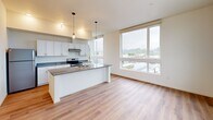 Located on the fifth floor at 310 Capitol Way N in downtown Olympia, Washington, this contemporary residence offers a bright and thoughtfully designed single-level layout. The open-concept living and kitchen area serves as the heart of the unit, featuring warm hardwood-style flooring that flows seamlessly throughout the space. The kitchen is outfitted with crisp white cabinetry, dark countertops, stainless steel appliances including a refrigerator and electric range, and a central island with pendant lighting overhead—ideal for both meal preparation and casual gatherings. Large floor-to-ceiling windows in the living area flood the space with natural light and frame sweeping views of Olympia's cityscape, waterway, and tree-lined hillsides beyond. The bedroom continues the clean, modern aesthetic with the same hardwood-style flooring, sage green accent walls, and oversized windows that offer expansive views of the surrounding urban and natural landscape. A wall-mounted HVAC unit provides individualized climate control. The overall ambiance of the unit is clean, airy, and modern, with an emphasis on natural light and elevated views that make it a standout offering in the heart of Olympia.