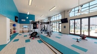 This well-appointed fitness center is designed to accommodate a wide range of workout routines within a single, expansive single-story space. The interior is immediately distinguished by its soaring ceilings and floor-to-ceiling windows along one side, which flood the facility with natural light and provide open views of an outdoor pool area and surrounding landscape. Large glass doors offer direct access to the exterior, further connecting the indoor and outdoor environments.

The design scheme is modern and energetic, featuring teal textured wall panels paired with crisp white walls and ceilings. The flooring consists of a patterned carpet tile in neutral tones accented by teal blocks, which visually define workout zones throughout the space. Multiple wall-mounted flat-screen televisions are positioned throughout the gym for convenience.

The fitness floor is comprehensively equipped, featuring cardio machines including treadmills, ellipticals, and stationary bikes, as well as a multi-station cable and strength machine setup, a punching bag, free weights, dumbbells, medicine balls, and battle ropes. A separate glass-enclosed room serves as a dedicated studio space for cycling or group fitness. Large ceiling fans provide additional airflow, and restroom access is available directly within the facility.