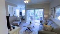 Located in Gulf Shores, Alabama, this single-floor space offers a welcoming and well-designed interior that blends comfort with functionality. The open-concept layout seamlessly connects the living and dining areas, creating a spacious and cohesive environment ideal for relaxing or entertaining.

The living area is anchored by a plush seating arrangement centered around a wooden coffee table and a patterned area rug, giving the space a warm, coastal character. A ceiling fan overhead provides both comfort and style. The dining area, positioned just adjacent, features a round table with ample seating and is bathed in natural light streaming through large windows fitted with blinds.

One of the standout features of this space is the expansive sliding glass door that opens to an outdoor patio or balcony area, flooding the interior with natural light and offering views of the lush greenery outside. Vertical blinds provide privacy and light control as needed.

The flooring throughout is a light-toned hardwood or wood-look surface that ties the rooms together cohesively. The neutral color palette — soft grays, whites, and warm wood tones — creates a bright and airy ambiance that is both modern and inviting, perfectly suited to its coastal Gulf Shores setting.