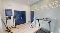 Located at 291 Alabaster Dr in Dothan, Alabama, this fitness space offers a focused and energizing environment designed for personal training and exercise. The single-floor layout is efficiently organized, connecting a main workout area with a hallway that leads to additional areas of the broader property.

The workout room immediately captures attention with its bold "Stronger Than Yesterday" wall lettering set against a soft sage green accent wall, complemented by a deep navy blue padded wall panel — together creating a motivating and visually dynamic atmosphere. Light wood-look flooring runs throughout, keeping the space feeling clean, bright, and modern.

The room is outfitted with a practical array of fitness equipment, including a NordicTrack treadmill, a stationary spin bike, a multi-function power tower and dip station, a weight bench, a dumbbell rack, and wall-mounted resistance band storage. A set of bold navy double entry doors and a frosted glass interior door lend the space a polished, intentional design aesthetic.

Natural light filters in through a window dressed with wood-toned blinds, adding warmth to the otherwise crisp white walls. Recessed ceiling lighting ensures the space remains well-lit. A posted fitness rules sign near the entrance indicates this room serves as a shared or community-use facility. Overall, the space is purpose-built, neatly appointed, and thoughtfully designed to support a consistent fitness routine.