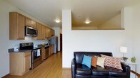This single-floor space is designed with an open-concept layout that seamlessly connects the kitchen and living areas, creating a cohesive and functional environment. The kitchen is well-appointed with warm honey-toned wood cabinetry that extends to the upper walls, providing generous storage. Stainless steel appliances, including a refrigerator, stove, and over-the-range microwave, lend a modern touch. A sink and countertop workspace complete the cooking area, all set atop light hardwood flooring that runs throughout the space.

A structural column subtly divides the kitchen from the living area, where a dark leather sofa accented with colorful throw pillows anchors the room. Flush-mount ceiling lights in both zones provide consistent, even illumination. The neutral white walls and light flooring contribute to a bright, clean aesthetic throughout.

The unit also features an outdoor balcony, offering views of a tree-lined neighborhood with nearby residential buildings visible in the distance — a pleasant outdoor extension of the living space. Overall, the layout is practical and comfortable, suited for everyday living.