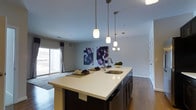 This single-floor residence offers a thoughtfully designed open-concept living space that seamlessly blends style and functionality. The kitchen serves as the centerpiece of the home, anchored by a generous island topped with sleek white countertops and equipped with an undermount sink and contemporary fixtures. Dark cabinetry provides a striking contrast to the light countertops, while a trio of pendant lights suspended above the island adds both ambiance and practical illumination. The open layout connects the kitchen directly to the living area, which features warm hardwood flooring that runs throughout the space, contributing to a cohesive and inviting atmosphere. A sliding glass door floods the living area with natural light and opens onto a private balcony, extending the living space outdoors. The walls are adorned with bold, abstract artwork featuring deep purple and black tones, adding a pop of color and personality to the otherwise neutral palette of white and grey tones. Recessed lighting in the ceiling complements the pendant fixtures, ensuring the space is well-lit at all times. The overall ambiance is modern and refined, with clean lines and a carefully curated aesthetic that makes this residence both functional and visually appealing.