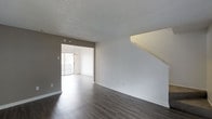 This two-storey space offers a clean, functional layout anchored by a spacious main living area on the ground floor. Dark hardwood-style flooring runs throughout, providing a strong visual contrast against the crisp white baseboards and neutral grey-toned walls. A textured popcorn ceiling adds subtle dimension overhead.

A staircase along the right wall connects the two floors, its open railing design contributing to an airy feel within the living space. The room flows naturally into an adjoining area, distinguished by light-toned flooring and a pendant light fixture overhead. A large sliding glass door in this secondary space allows generous natural light to pour in, brightening the overall interior and providing outdoor access.

The neutral color palette throughout — combining soft grey walls, white trim, and contrasting flooring — creates a versatile backdrop suitable for a range of furnishing styles. The layout is straightforward and practical, with clearly defined zones between the main living area and the lighter adjoining room. Overall, the space presents a tidy, move-in ready environment with a simple, modern aesthetic across both levels.