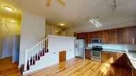 This thoughtfully designed single-floor space combines functionality and style through an open-concept layout centered around gleaming natural hardwood floors that run throughout the main living areas. The spacious living room offers a generous, airy feel, accented by a ceiling fan and track lighting that illuminate the room effectively. A staircase with a classic white banister leads up to an elevated loft area, adding an architectural dimension and additional usable space to the unit.

The kitchen is a standout feature, offering rich warm-toned wood cabinetry that extends from floor to ceiling, providing ample storage. Granite countertops in a speckled neutral tone offer a polished contrast to the cabinetry. A full suite of stainless steel appliances — including a refrigerator with ice maker, electric range, built-in microwave, and dishwasher — ensures a modern and practical cooking experience. A row of large windows fitted with horizontal blinds floods the kitchen with natural light while maintaining privacy.

The bathroom is clean and functional, featuring tile flooring, a granite-topped vanity, and a full bathtub with shower enclosure. White cabinetry provides additional storage, completing a well-organized and practical space. Overall, the unit presents a cohesive, comfortable environment suited for everyday living.