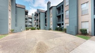This multi-story residential complex presents a cohesive architectural design characterized by distinctive teal-green wood siding and neutral stucco accents. The buildings rise several stories, featuring prominent vertical chimney structures and pitched rooflines that add visual interest to the exterior profile. Each level is equipped with private balconies framed by matching teal railings, offering residents their own outdoor space. The complex is arranged in a courtyard-style layout, with a generously sized concrete common area at its center serving as a shared gathering and circulation space. Manicured hedgerows line portions of the exterior, providing a degree of greenery and definition between buildings. Wooden privacy fencing in a coordinating teal tone delineates boundaries within the property. Large windows are distributed across the facades, allowing natural light into individual units. The overall aesthetic is consistent and well-maintained, with a cohesive color palette unifying the multiple structures. The open courtyard design promotes a sense of community while maintaining individual privacy through strategic building placement and landscaping.