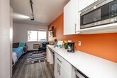 Located at 4117 University Ave in Cedar Falls, Iowa, this efficiently designed studio unit makes excellent use of its compact footprint. The open-plan layout seamlessly integrates a living area and kitchenette into a single, well-organized space. A standout feature is the built-in Murphy bed, which folds neatly against the wall to free up floor space during the day, offering flexibility without sacrificing comfort.

The kitchenette is outfitted with white cabinetry, a countertop workspace, an under-counter refrigerator, and a microwave — providing all the essentials for everyday living. Track lighting along the ceiling adds a modern touch while ensuring the space is well-lit throughout.

The living area is highlighted by a bold orange accent wall, a wall-mounted flat-screen TV, and a window that invites natural light and outdoor views. Wood-look plank flooring runs throughout, adding warmth and continuity, while a patterned area rug anchors the seating zone.

The bathroom features a walk-in shower, a vanity with white cabinetry and an integrated sink, and a large mirror — clean and functional in design.

Overall, the unit's bright color palette, smart storage solutions, and thoughtful layout create a practical and inviting living environment suited for individuals seeking efficient, all-in-one accommodation in Cedar Falls.