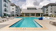 Located at 49 N Assembly Dr in Wilmington, NC, this two-story residential community showcases a thoughtfully designed coastal-inspired architectural style. The exterior features a combination of light gray and blue board-and-batten siding, white trim, metal standing-seam roofing, and black iron balcony railings that lend a clean, contemporary coastal aesthetic to the multi-building complex.

At the heart of the community lies a generously sized resort-style swimming pool with crystal-clear blue water, complemented by a shallow lounge shelf ideal for relaxing. The expansive pool deck is outfitted with a generous collection of chaise lounge chairs, shaded by large white market umbrellas, and accented with small side tables for a complete outdoor retreat experience. A dedicated clubhouse structure with large glass-panel walls and a covered outdoor porch area overlooks the pool, serving as a central amenity hub.

Surrounding the pool and throughout the courtyard, professionally curated landscaping features ornamental grasses, flowering shrubs, and mulched garden beds that add vibrant greenery and color. Paved walkways connect the various outdoor areas, including additional lounge seating zones and intimate courtyard nooks. Each residential building features private balconies on upper floors, offering outdoor space and views over the beautifully landscaped grounds below.