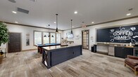 Located in Lawrenceville, Georgia, this game room offers a generously sized, single-floor recreational space designed for both entertainment and casual gatherings. The layout is open and thoughtfully arranged, anchored by a full-size pool table with a vibrant blue felt surface, lit from above by a cluster of industrial-style pendant lights that lend the room a modern farmhouse character.

A standout feature is the dark navy, X-base communal island that serves as a coffee bar, complete with bar-height seating and a chalkboard menu wall behind it — creating an inviting social hub within the space. Recessed lighting throughout the ceiling complements the pendant fixtures, ensuring the room is well-lit at all times.

Large windows line the walls, flooding the interior with abundant natural light and offering views of the surrounding greenery. The neutral-toned carpet adds warmth and texture underfoot, tying together the earthy, relaxed aesthetic of the room. A comfortable leather lounge chair and side table occupy one corner, providing a quiet spot to relax.

Decorative touches including framed artwork, potted plants, and tasteful accents contribute to the polished yet approachable ambiance. A connected hallway provides access to and from the space. Overall, this game room presents a versatile, welcoming environment suited for recreation and community engagement.