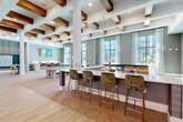 Located in Allen, Texas, this thoughtfully designed community amenity space delivers a modern, upscale atmosphere across a single open floor. The standout architectural feature is the striking coffered ceiling adorned with rich wood beams, complemented by brass pendant chandeliers that cast a warm, sophisticated glow throughout the space. Light hardwood-style flooring transitions seamlessly into patterned carpeted areas, delineating distinct activity zones while maintaining a cohesive aesthetic.

The lounge and gaming area is well-equipped, featuring a pool table, a foosball table, and a large-format display screen mounted against a textured stone accent wall — ideal for social gatherings. Velvet and leather upholstered seating in warm amber and olive tones is arranged throughout, offering comfortable spots to relax or socialize. Multiple bar-height counters with stylish gold-legged stools provide additional seating and workspace options.

Floor-to-ceiling windows and glass doors line the exterior walls, flooding the interior with abundant natural light and offering unobstructed views of the outdoor pool area and surrounding urban landscape. A dedicated work or co-working nook near the entrance features a wall-mounted TV and compact seating. The space's teal and gray accent walls, curated artwork, and layered lighting contribute to a vibrant yet refined community environment.