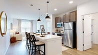 Located at 4455 Condor Dr in Hilliard, Ohio, this thoughtfully designed one-bedroom, one-bathroom residence offers a clean, modern aesthetic across a single floor. The open-concept layout seamlessly connects the kitchen and living room, creating an inviting and functional living space. The kitchen is a standout feature, showcasing warm-toned cabinetry, a large island with a white quartz countertop and undermount sink, stainless steel appliances, and bold matte black pendant lighting overhead. Light wood-style flooring flows throughout, enhancing the bright, airy feel of the space. The living area is generously proportioned, bathed in natural light from large windows dressed with linen curtains and slatted blinds, offering views of the surrounding greenery. The bedroom is a comfortable retreat featuring dual windows, ample natural light, and direct access to the bathroom. The bathroom includes a wood-vanity with a white countertop, a tiled tub-shower combination, and black hardware accents. A dedicated closet provides practical storage. The overall ambiance is contemporary and neutral, with warm wood tones and crisp white walls creating a welcoming, well-balanced interior throughout.