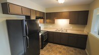 This single-floor space includes a kitchen designed for practical use, showcasing dark wood cabinetry that lines the upper and lower walls, providing generous storage throughout. The kitchen is equipped with black appliances, including a full-sized refrigerator, a gas range with an overhead exhaust hood, and a built-in microwave. A double-basin stainless steel sink is positioned along the far wall beneath additional upper cabinetry, complemented by light-toned countertops that contrast with the darker cabinet finish. The flooring features earthy, stone-look tile laid in a natural pattern, adding texture and visual warmth to the space. Overhead, a flush-mount ceiling light provides consistent illumination throughout the kitchen area. The layout follows an L-shaped configuration, allowing for efficient workflow and clear countertop workspace. The light-colored walls offer a neutral backdrop that balances the richness of the dark cabinetry, creating a cohesive and functional kitchen environment suited for everyday cooking and meal preparation.
