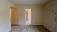 This single-floor space presents a straightforward and functional layout characterized by neutral tones throughout. The interior features wall-to-wall carpeting in a light beige tone, contributing to a quiet and understated ambiance. The walls are finished in a clean off-white, creating a bright and open feel despite the absence of visible windows in the primary view. Standard ceiling heights provide a comfortable sense of space without excess volume. A doorway connects the main room to an adjacent space, which appears to feature a louvered bifold closet door, suggesting additional storage within the layout. The primary room also includes a door along the left wall, offering further access or enclosure options. Electrical outlets are positioned at standard heights along the walls. The overall design is minimal and utilitarian, providing a blank canvas that lends itself well to personalization. The connected rooms share consistent finishes, maintaining a cohesive aesthetic throughout the space. The layout appears practical and efficient, suited for everyday residential use.