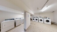This shared laundry facility offers a practical and well-organized space designed to accommodate multiple users efficiently. The room is laid out to maximize functionality, featuring a generous arrangement of front-loading washing machines and top-loading dryers positioned along the walls and around a central structural column that divides the space into distinct zones. Each machine is equipped with a digital payment interface, providing a convenient and modern approach to laundry management. The flooring throughout is durable terrazzo, offering a clean and easy-to-maintain surface that complements the room's utilitarian aesthetic. Walls are finished in crisp white, contributing to a bright and hygienic atmosphere, further enhanced by fluorescent lighting mounted to the ceiling. A small window along one wall allows for a modest amount of natural light. The space also includes visible plumbing connections and utility hookups integrated neatly along the walls, as well as a fire extinguisher for safety compliance. A notice board is mounted on the central column, likely displaying usage guidelines and policies. Access is provided through a solid entry door fitted with a window panel and ventilation louver. Overall, the facility is clean, functional, and well-maintained, catering effectively to the laundry needs of a shared residential building.