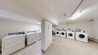This shared laundry facility offers a practical and well-organized space designed to accommodate multiple users efficiently. The room is laid out to maximize functionality, featuring a generous arrangement of front-loading washing machines and top-loading dryers positioned along the walls and around a central structural column that divides the space into distinct zones. Each machine is equipped with a digital payment interface, providing a convenient and modern approach to laundry management. The flooring throughout is durable terrazzo, offering a clean and easy-to-maintain surface that complements the room's utilitarian aesthetic. Walls are finished in crisp white, contributing to a bright and hygienic atmosphere, further enhanced by fluorescent lighting mounted to the ceiling. A small window along one wall allows for a modest amount of natural light. The space also includes visible plumbing connections and utility hookups integrated neatly along the walls, as well as a fire extinguisher for safety compliance. A notice board is mounted on the central column, likely displaying usage guidelines and policies. Access is provided through a solid entry door fitted with a window panel and ventilation louver. Overall, the facility is clean, functional, and well-maintained, catering effectively to the laundry needs of a shared residential building.