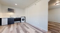 This single-floor unit offers a clean, modern aesthetic defined by light-toned vinyl plank flooring that runs consistently throughout every room, contributing to a cohesive and well-maintained interior. The layout includes a main living area with multiple windows fitted with blinds, allowing for natural light while maintaining privacy. A dedicated entry area features a solid white door alongside additional window access, creating a bright and welcoming entryway.

The kitchenette is equipped with dark cabinetry, a sink, and an in-unit washer, providing essential convenience within a compact footprint. Exposed piping along the ceiling in this area adds an industrial accent to the otherwise clean design. A wall-mounted HVAC unit is present in one of the rooms, ensuring climate control, accompanied by a window and track-style ceiling rails.

The bathroom is thoughtfully designed with ADA accessibility in mind, featuring stainless steel grab bars positioned around the toilet, a window for natural ventilation, and a floating vanity with a dark base. The neutral white walls throughout the unit create a bright, open atmosphere. The 3D model reveals an arched interior doorway, adding a subtle architectural detail to the otherwise straightforward floor plan. Overall, the space is functional, accessible, and move-in ready.
