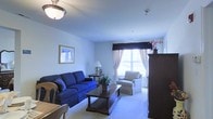 This single-floor space centers around a comfortable and thoughtfully arranged living room. The room features light-colored walls and soft, wall-to-wall carpeting that contributes to a warm and inviting atmosphere. A large window dressed with sheer curtains and a dark decorative valance allows natural light to fill the space, brightening the interior considerably.

The living area is furnished with a bold navy blue sofa accented by coordinating throw pillows, paired with a neutral-toned upholstered armchair positioned near the window. A dark wood coffee table serves as a central anchor between the seating pieces, complemented by a matching side table and a traditional-style table lamp. A tall dark wood armoire stands along one wall, providing both storage and visual contrast against the lighter surroundings.

Wall décor includes a framed artwork piece that adds a touch of character to the space. Overhead, a flush-mount ceiling light fixture provides consistent illumination throughout the room. A small potted plant near the coffee table introduces a subtle natural element. Overall, the space presents a classic, well-maintained aesthetic suitable for comfortable everyday living.