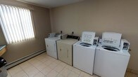 This single-floor laundry facility is designed with practicality and convenience in mind. The room is equipped with multiple top-loading washing machines and front-loading dryers arranged side by side along the wall, offering ample capacity for shared use. The light beige tile flooring is easy to clean and maintain, contributing to the room's functional character. Neutral-toned walls give the space a clean, understated appearance, while a window along one side allows natural light to brighten the interior and create a more welcoming atmosphere. A baseboard heater runs along the base of the wall beneath the window, ensuring the room remains comfortable year-round. Each machine features coin-operated or card-operated controls, making the facility straightforward to use. The overall layout is open and uncluttered, allowing users to move freely between machines. This laundry room serves as a well-organized, no-frills shared amenity that prioritizes ease of use and accessibility.