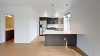 This thoughtfully designed single-floor unit showcases a contemporary aesthetic throughout, with light hardwood flooring and crisp white walls creating a clean, cohesive look from room to room. The open-concept kitchen serves as the heart of the space, anchored by a large central island with a white quartz countertop, an integrated sink, and pendant lighting overhead. Dark gray cabinetry pairs with white subway tile backsplash, open wood shelving, and a full suite of stainless steel appliances — including a side-by-side refrigerator with ice and water dispenser, an electric range, and an over-the-range microwave. A window above the sink brings in natural light and outdoor views. The main living area is bright and open, featuring a large window that frames views of mature trees, allowing generous sunlight to pour across the hardwood floors. A wall-mounted HVAC unit provides climate control. The unit includes two bathrooms, both finished with gray vanity cabinetry, white countertops, and large mirrors. One bathroom features a glass-enclosed walk-in shower with white subway tile surround, while the second offers a more compact layout. Both bathrooms continue the hardwood flooring found throughout the rest of the unit, maintaining a seamless, modern finish.
