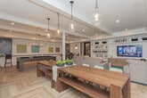 Located in Brookhaven, Georgia, this single-floor community amenity space at 3519 Sunderland Way NE offers a thoughtfully designed environment for socializing and relaxation. The interior is anchored by light herringbone-patterned hardwood flooring and tray ceilings with recessed lighting throughout, lending a polished, contemporary feel to the entire space.

The main lounge area features multiple seating arrangements, including plush sofas and modern accent chairs arranged around a large flat-screen TV framed by expansive built-in bookshelves — ideal for both casual gatherings and quiet leisure. A set of French doors floods the space with natural light and provides seamless access to the outdoor pool area, visible through the glass.

A dedicated entertainment zone houses a full-sized billiards table, complemented by additional lounge seating and support columns that add architectural character. The kitchenette is well-appointed with warm wood cabinetry, open industrial-style shelving, white subway tile backsplash, stainless steel appliances, and a butcher block communal dining table illuminated by globe pendant lights. The overall ambiance is modern yet inviting, blending natural materials with clean lines to create a versatile and comfortable community hub.