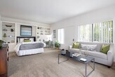 Located at 1142 Buckingham Dr in Costa Mesa, California, this single-floor residence offers a clean, functional layout with a light and airy aesthetic throughout. The living room is a welcoming space featuring plush wall-to-wall carpeting, neutral walls, and multiple large windows dressed with vertical blinds that allow generous natural light to filter in. The open, relaxed layout comfortably accommodates seating arrangements and provides a bright, pleasant atmosphere. Adjacent to the living area, the galley-style kitchen is efficiently designed with ample white cabinetry, granite-toned countertops, a double-basin sink, hardwood-style flooring, and white appliances including a refrigerator and range. A breakfast bar connects the kitchen to the broader living space, enhancing the open feel. The bedroom is generously proportioned with soft carpeting and open sightlines to both the kitchen and a built-in shelving area. A wall-mounted TV and dedicated storage space add to the room's practicality. The bathroom is well-appointed with a granite-topped vanity, a large wall mirror, a combination tub and shower, and warm wood-style flooring. Throughout the residence, the consistent use of neutral tones and natural light creates a cohesive, comfortable living environment ideal for everyday living in Southern California.