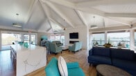 This single-story communal space showcases a coastal-contemporary design defined by striking vaulted ceilings with exposed white-painted beams and shiplap detailing that lend the interior a bright, airy character. Herringbone-patterned wood-look flooring runs throughout, providing a cohesive visual foundation across all areas.

The main lounge area is generously proportioned, featuring multiple seating arrangements anchored by a wall-mounted flat-screen television above a sleek linear electric fireplace. Furnishings in navy blue, teal, and patterned aqua tones reinforce the coastal aesthetic. Large windows and glass doors frame outdoor views and flood the interior with natural light.

A dedicated dining nook, positioned near full-length glass doors opening to an exterior walkway, accommodates a round table with upholstered seating. Adjacent to the main lounge, a full kitchen includes stainless steel appliances, taupe shaker-style cabinetry, a tile backsplash, and a large quartz-topped island with bar seating — suitable for events and gatherings.

Artwork with nautical and botanical themes accents the walls, complemented by statement lighting fixtures and a textured circular mirror. The space benefits from multiple points of outdoor access, reinforcing a seamless indoor-outdoor flow throughout.