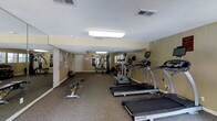 This fitness center offers a comprehensive workout environment designed to accommodate a wide range of exercise routines. The space is outfitted with multiple SportsArt treadmills positioned along one side, providing dedicated cardio zones for users. A stationary bike adds further cardiovascular options, while a multi-function cable and weight machine anchors the strength training area toward the back of the room. A workout bench and free weights are also available, rounding out the resistance training offerings. A stability ball and additional accessories are neatly placed throughout the space, contributing to a versatile training environment. The room features neutral yellow-beige walls, a textured ceiling with flush-mounted fluorescent lighting that keeps the interior well-lit, and durable low-pile carpeted flooring suited for gym use. Wall-mounted mirrors near the entrance enhance the sense of space and allow users to monitor their form. A door at the rear provides additional access or storage connectivity. Informational postings on the walls offer guidance for users. The layout is open and functional, allowing comfortable movement between equipment stations, making this fitness center a practical and fully equipped space for daily exercise needs.