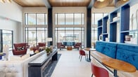 Located in Huntsville, AL, this elegant lobby and lounge space delivers a striking first impression through its modern architectural design and thoughtfully curated interiors. The entrance foyer welcomes visitors with soaring double-height ceilings, a dramatic tiered chandelier, large-format tile flooring, and a warm wood accent wall paired with a statement fireplace. A bold, navy-clad fireplace column rises to the ceiling in the main lounge area, anchoring a comfortable seating arrangement complete with a tufted sofa, swivel armchairs, and a sculptural pendant light fixture. The open-concept layout flows seamlessly into a bright social lounge, where floor-to-ceiling black-framed windows and glass walls flood the space with natural light and offer expansive outdoor views. A vibrant blue built-in shelving unit serves as a stylish focal point alongside banquette booth seating and café-style tables, creating an inviting area for casual gatherings. A warm wood-paneled ceiling with exposed dark beams adds texture and architectural character overhead. Throughout, the design palette blends deep navy, rich burgundy, and warm wood tones to create a contemporary yet welcoming ambiance that is both functional and visually refined.