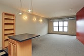 This single-floor unit offers a thoughtfully arranged open layout that combines living and kitchen spaces into one cohesive area. The main living space is carpeted in a neutral gray and anchored by a striking floor-to-ceiling wood-panel accent wall finished in warm orange-brown tones, adding a bold architectural focal point. Exposed ductwork runs along the upper perimeter, lending an industrial character to the space. A bank of large windows along one wall invites natural light and provides views of the surrounding urban streetscape.

The kitchen transitions seamlessly from the living area via a checkered tile floor, creating a clear visual boundary between the two zones. It is equipped with black appliances including a refrigerator, gas range, built-in microwave, and dishwasher, complemented by light wood cabinetry, dark countertops, and a double-basin sink. Open shelving and a small kitchen island add functional storage and workspace. Pendant lighting overhead illuminates the area warmly.

The bathroom features a classic black-and-white checkered tile floor, a vanity with a dark countertop, an under-mount sink, light wood cabinetry, and a large mirror with overhead lighting, completing the unit's practical and distinct aesthetic.