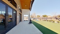 This single-level outdoor fitness space combines a covered concrete workout area with an expansive artificial turf field, designed to support a wide range of functional training activities. The building facade features a modern aesthetic with warm wood-paneled siding, board-and-batten white accents, and stone column bases, giving the space a contemporary yet inviting character. A sloped wood-planked ceiling extends over the covered portion, providing shade while maintaining an open-air feel.

Large glass sliding doors and floor-to-ceiling windows connect the covered outdoor zone to an interior fitness facility, allowing visual continuity between the two spaces. The covered concrete pad is equipped with functional training tools including kettlebells, weighted sleds, weight plate racks, and a push/pull sled, making it well-suited for high-intensity workouts. The turf field, which extends generously beyond the covered area, features agility ladders laid out for speed and coordination drills, along with additional portable training equipment.

The entire area is enclosed by a sleek black iron fence, with low landscaping plantings along the perimeter adding a polished, finished appearance. Open sky views and abundant natural light contribute to a motivating and energetic training environment.