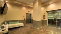 This single-floor space presents a functional and well-organized lobby or common area with a straightforward, utilitarian design aesthetic. The interior is finished with warm beige and tan wall tones accented by a two-tone treatment, with a darker wainscoting-style lower panel running along the perimeter walls, adding a subtle architectural detail that grounds the space visually.

A stainless steel elevator with standard call panel is centrally positioned, serving as a key functional element of the area. The flooring is a polished concrete surface that adds an industrial character to the otherwise neutral palette. A small entry mat is placed in front of the elevator for practical use.

Recessed lighting provides even, ambient illumination throughout the space. A modest seating area is present, featuring a low white bench with a sage green back panel, offering a simple resting spot for occupants. Safety and utility features are visible on the walls, including a mounted fire safety cabinet and a communication panel, reflecting the space's practical, institutional character. Structural columns break up the open floor plan, contributing to the overall layout and flow of the area.