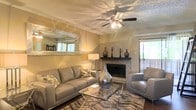 This single-floor space centers around a well-appointed living room that combines comfort and style. The room is anchored by a classic brick fireplace with a dark wood mantel, currently styled with decorative vases and candle holders, serving as a natural focal point. A ceiling fan with an integrated light fixture is mounted on the textured ceiling, providing both airflow and ambient lighting throughout the space.

The living area is furnished with a contemporary grey leather sofa and matching accent chair, arranged around a glass-top coffee table atop a patterned area rug, creating a cohesive and inviting seating arrangement. A large decorative mirror with a distressed white frame adorns one wall, adding depth and reflecting natural light into the room. Warm golden-toned walls contribute to the overall inviting ambiance.

Natural light filters in through a sliding glass door on one side, offering a view of an outdoor balcony or patio area with greenery visible beyond. A secondary window on the adjacent wall further enhances the brightness of the space. Dark hardwood flooring grounds the room and provides a pleasing contrast to the lighter furnishings and wall tones.