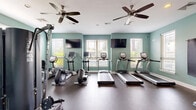 Located at 475 Swanholme Dr in Murfreesboro, Tennessee, this fitness center offers a thoughtfully designed exercise space spread across a single floor. The room is finished with soft seafoam-green walls and durable rubber flooring, creating a clean and energizing environment well-suited for a variety of workouts.

The cardio area is well-appointed with multiple treadmills and elliptical machines, each positioned near windows fitted with blinds that allow natural light to filter in while providing privacy. Wall-mounted flat-screen televisions are positioned for easy viewing during cardio sessions. A ceiling fan overhead contributes to air circulation and comfort throughout the room.

On the strength training side, the space includes a multi-functional cable machine, an adjustable weight bench, and a dumbbell rack with a range of free weights. A large wall mirror spans a significant portion of the room, enhancing the sense of openness and allowing users to monitor their form. A yoga or exercise mat mounted on the wall adds versatility for stretching or floor-based workouts.

The space is accessed through a glass-paneled door and is equipped with standard safety features. Recessed and overhead lighting, combined with ample natural light from several windows, ensures the room remains bright and welcoming throughout the day.