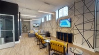 This contemporary single-floor communal lounge is designed with a sharp, modern aesthetic that balances comfort and visual impact. The most striking architectural feature is a large-scale geometric line mural that dominates one wall, rendered in bold black lines against a neutral background, creating a dynamic focal point for the space. Multiple wall-mounted flat-screen televisions are positioned throughout the lounge, ensuring clear sightlines from every seating area.

The furniture arrangement is both stylish and functional, featuring plush gray sectional sofas adorned with patterned throw pillows, complemented by vibrant yellow accent chairs with sleek metal frames. Small round glass and metal side tables are distributed throughout, providing convenient surfaces for guests. The flooring consists of warm-toned wood-look planks that contrast elegantly with the darker architectural column accents finished in charcoal.

Lighting is delivered through a combination of linear LED ceiling fixtures and subtle wall sconces, creating an even, well-lit environment. High clerestory windows along one wall allow natural light to filter in while maintaining privacy. A glass-paneled door connects this lounge to an adjacent space, enhancing the open flow of the layout. Open shelving with decorative objects adds a refined, curated touch to the overall design.