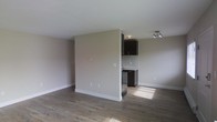 This single-floor space offers a clean, contemporary design characterized by an open and airy layout. The main living area is finished with rich, dark hardwood-style flooring that contrasts elegantly with the soft, neutral-toned walls and crisp white baseboards and ceiling, creating a bright and welcoming ambiance. Natural light filters into the space, enhancing its open feel. A half-wall partition subtly defines the kitchen area from the main living space while maintaining a sense of openness throughout. The kitchen nook is appointed with sleek dark cabinetry, a matching dark appliance suite, and light countertops, offering a stylish contrast to the surrounding neutral tones. Modern track lighting fixtures are mounted to the ceiling, providing well-distributed illumination across both the kitchen and living areas. The overall design is minimalist and functional, making efficient use of the available space. The consistent flooring running throughout ties the areas together seamlessly, while the restrained color scheme gives the space a versatile, move-in-ready quality suited to a variety of interior styles.