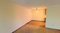 This single-story space offers a clean, functional layout with a warm and neutral aesthetic. The main living area is finished with wall-to-wall carpet, providing a comfortable and quiet underfoot feel, while an adjacent section transitions to hardwood-style flooring, adding visual variety and practicality to the space. The walls are painted in a soft, off-white tone that reflects light and contributes to an open, airy atmosphere.

A ceiling fan with an integrated light fixture is centrally positioned, serving both as a functional lighting source and an aid for air circulation throughout the space. The baseboards are finished in a warm wood tone, complementing the flooring and adding a cohesive design element throughout the room.

A glimpse into the adjacent area suggests additional rooms beyond, with cabinetry visible in the background, hinting at a kitchen or utility space nearby. The overall layout flows naturally from one area to the next, making the space feel connected and easy to navigate. The simple, understated design makes this space highly adaptable to a variety of furnishing styles and functional uses.