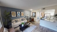 This single-floor residence offers a thoughtfully designed open-concept layout that seamlessly connects the living, dining, and kitchen areas. The living room is a comfortable and stylish space, highlighted by rich wood-look flooring, a textured ceiling, and a striking accent wall in a cool slate blue. A large window fitted with layered blinds and draping curtains invites natural light while maintaining privacy, and the space accommodates a full seating arrangement with room for a cozy reading nook near the entry. A trio of geode-inspired artwork adds a distinctive decorative touch to the main wall. The dining area transitions naturally from the living space, providing a compact yet functional setting for meals. The galley-style kitchen is efficiently organized with ample white upper and lower cabinetry, stone-look countertops, a stainless steel undermount sink, and a full suite of stainless steel appliances including a refrigerator and range. A skylight-style overhead fixture brightens the kitchen throughout the day. The bathroom features a tub and shower combination enclosed by a curved rod and clean white curtain, keeping the space bright and functional. Throughout the residence, consistent wood-look flooring and a neutral color palette create a cohesive, modern ambiance.