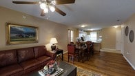 This single-floor home offers a seamlessly connected living, dining, and kitchen layout that creates an open and functional living environment. The living area is anchored by rich hardwood flooring and features a classic ceiling fan with light fixture overhead, complemented by a textured ceiling. A deep brown leather sofa, an ornate framed painting, and a decorative area rug contribute to the warm, traditional aesthetic of the space.

The dining area flows naturally from the living room, centered around a dark wood dining table with upholstered red accent chairs, providing ample seating for gatherings. A table lamp on a black side console adds a soft, ambient glow to the transitional space between the living and dining areas.

The kitchen is positioned at the far end of the open floor plan, benefiting from natural light streaming through curtained windows. Light wood cabinetry, a white refrigerator, and a functional countertop workspace define the kitchen's layout, which also includes a peninsula that connects it to the dining area.

Throughout the home, warm beige-toned walls tie each area together cohesively, while the consistent hardwood flooring enhances the sense of flow and continuity. The overall ambiance is cozy, traditional, and well-suited for comfortable everyday living.