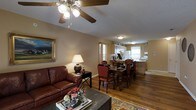 This single-floor home offers a seamlessly connected living, dining, and kitchen layout that creates an open and functional living environment. The living area is anchored by rich hardwood flooring and features a classic ceiling fan with light fixture overhead, complemented by a textured ceiling. A deep brown leather sofa, an ornate framed painting, and a decorative area rug contribute to the warm, traditional aesthetic of the space.

The dining area flows naturally from the living room, centered around a dark wood dining table with upholstered red accent chairs, providing ample seating for gatherings. A table lamp on a black side console adds a soft, ambient glow to the transitional space between the living and dining areas.

The kitchen is positioned at the far end of the open floor plan, benefiting from natural light streaming through curtained windows. Light wood cabinetry, a white refrigerator, and a functional countertop workspace define the kitchen's layout, which also includes a peninsula that connects it to the dining area.

Throughout the home, warm beige-toned walls tie each area together cohesively, while the consistent hardwood flooring enhances the sense of flow and continuity. The overall ambiance is cozy, traditional, and well-suited for comfortable everyday living.