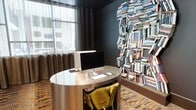 This single-floor space makes an immediate impression with its bold, contemporary design and thoughtfully curated details. The standout feature is a dramatic wall installation composed entirely of stacked books, artfully arranged in the silhouette of a human head — a striking piece that serves as both art and a nod to intellect and creativity. Dark, moody walls in deep charcoal provide a sophisticated backdrop that allows this installation to command attention.

The workspace is anchored by a sleek, oval metallic desk with a polished reflective surface, paired with a plush mustard-yellow accent chair that introduces a vibrant pop of color against the darker palette. An iMac desktop computer sits atop the desk, reinforcing the space's function as a productive yet design-forward work area.

Natural light filters through a large window dressed with floor-length drape curtains in warm brown tones, softening the room's darker aesthetic and lending an airy quality to the interior. The flooring features a patterned carpet in warm, earthy tones that complements the overall color scheme.

The result is a space that balances functionality with artistic expression — an environment designed to inspire focus and creativity in equal measure.