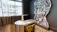 This single-floor space makes an immediate impression with its bold, contemporary design and thoughtfully curated details. The standout feature is a dramatic wall installation composed entirely of stacked books, artfully arranged in the silhouette of a human head — a striking piece that serves as both art and a nod to intellect and creativity. Dark, moody walls in deep charcoal provide a sophisticated backdrop that allows this installation to command attention.

The workspace is anchored by a sleek, oval metallic desk with a polished reflective surface, paired with a plush mustard-yellow accent chair that introduces a vibrant pop of color against the darker palette. An iMac desktop computer sits atop the desk, reinforcing the space's function as a productive yet design-forward work area.

Natural light filters through a large window dressed with floor-length drape curtains in warm brown tones, softening the room's darker aesthetic and lending an airy quality to the interior. The flooring features a patterned carpet in warm, earthy tones that complements the overall color scheme.

The result is a space that balances functionality with artistic expression — an environment designed to inspire focus and creativity in equal measure.
