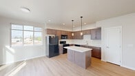 Located at 910 E Fourth Ave in Post Falls, Idaho, this single-floor unit offers a thoughtfully designed open-concept layout. The kitchen is a standout feature, showcasing rich dark wood cabinetry, a central island with a white countertop, pendant lighting, and black appliances including a refrigerator, range, and built-in microwave. Light wood-style flooring flows throughout the main living area, which is bathed in natural light from large windows and sliding glass doors that open to a private balcony with views of the surrounding landscape and mountains. A wall-mounted mini-split unit provides efficient climate control in the living space, which also includes an in-unit laundry area. The two bedrooms feature plush carpeting and windows that bring in ample natural light. Both bathrooms are finished with clean, modern fixtures, including tub/shower combinations, and one includes a vanity with a white countertop and a large mirror with contemporary lighting. The unit's neutral color palette and clean finishes create a bright, cohesive atmosphere throughout.