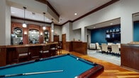 The Savoy Clubhouse in Chamblee, Georgia is a single-floor community facility designed with a sophisticated, upscale aesthetic that blends classic architectural details with modern comfort. Rich hardwood flooring flows throughout the main areas, complemented by dark wood wainscoting, crown molding, and bold wood trim that frame the space with a warm, refined character.

The central gathering area is anchored by a full-sized billiards table beneath an elegant chandelier, with arched windows and French doors that invite natural light and offer views of the lush outdoor surroundings. Adjacent to this space is a stylish bar area outfitted with dark cabinetry, black granite countertops, bar seating, pendant lighting, and mirrored backdrops that lend a classic lounge atmosphere.

A private dining or conference room, accented by a deep teal feature wall bearing the "SAVOY" name, provides an intimate setting with a large wood dining table and upholstered seating — ideal for gatherings or meetings. The business center features a workstation and is highlighted by a dramatic arched window framing verdant outdoor views. Additional cabinetry and a utility sink complete the practical amenities. Together, these spaces create a clubhouse that is both functional and distinguished.