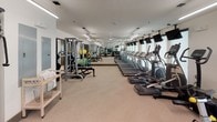 This fitness center offers a comprehensive workout environment designed to accommodate a wide range of exercise needs. The space is laid out across a single floor, with a clean, modern aesthetic defined by white walls, light-toned flooring, and crisp linear lighting suspended from the ceiling that ensures the entire area is evenly and brightly illuminated.

The cardio zone lines one side of the room and features a generous row of treadmills and elliptical trainers, each equipped with individual monitor displays mounted above for entertainment during workouts. A selection of stationary and spin bikes adds further variety for low-impact cardiovascular training.

The strength training section is well-appointed, offering a full dumbbell rack spanning a broad range of weights, multiple weight benches, a Smith machine, and a cable resistance system suitable for a variety of functional exercises. Medicine balls and additional free weights round out the functional fitness offerings.

Full-length mirrors run along one wall, visually expanding the space while allowing users to monitor their form throughout their workouts. Large windows with roller shades bring in natural light and contribute to the open, airy feel of the room. A wall-mounted television and additional ceiling fans enhance the overall comfort. The space is thoughtfully organized, providing ample room to move freely between stations, making it a practical and inviting facility for fitness enthusiasts of all levels.
