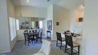 This single-story space showcases an open-concept layout that seamlessly connects a formal dining area with a bar-height counter seating section. The vaulted ceilings create a striking sense of vertical space, enhancing the overall feeling of openness throughout. Neutral-toned walls and light carpet flooring provide a clean, versatile backdrop that complements the bold contrast of dark wood furnishings.

The dining area is anchored by a round dark wood table surrounded by matching ladder-back chairs with dark upholstered seats. A large mirror on the adjacent wall reflects light and amplifies the sense of depth in the room. Decorative wall art, including a framed elephant print and a smaller accent piece, adds a curated, sophisticated touch to the space.

Adjacent to the dining area, a bar-height counter is lined with tall, dark wood bar stools featuring cream cushioned seats, offering an additional casual dining or entertaining option. A bowl of fresh fruit serves as a subtle decorative accent on the counter. A small console table with a decorative lamp is positioned nearby, contributing to the refined ambiance. Natural light filters in from a window visible in the background, brightening the overall atmosphere.